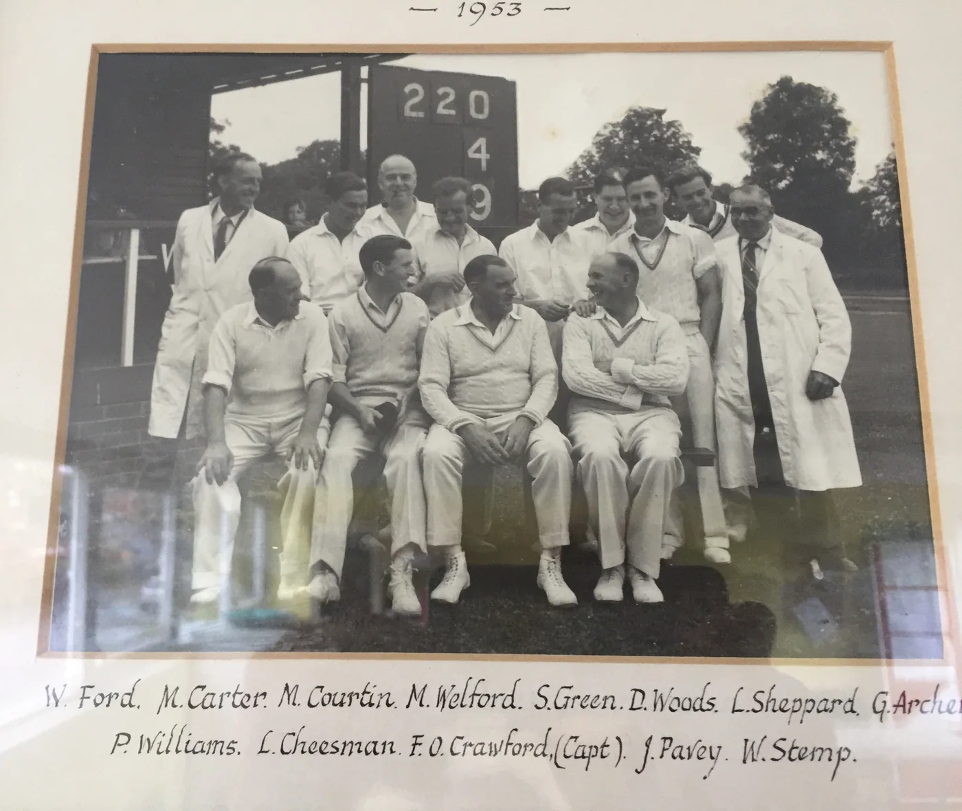 The 1953 Wisborough Green cricket team in whites, posed in front of the scoreboard reading 220 for 4